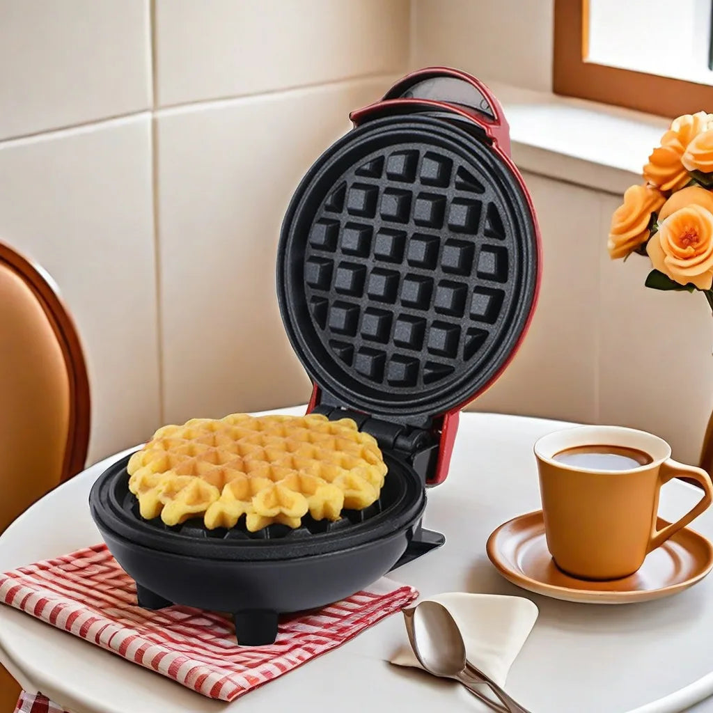 Red Versatile Mini Waffle Maker in use, cooking a waffle on a table with coffee, a spoon, and flowers for a cozy breakfast setup.