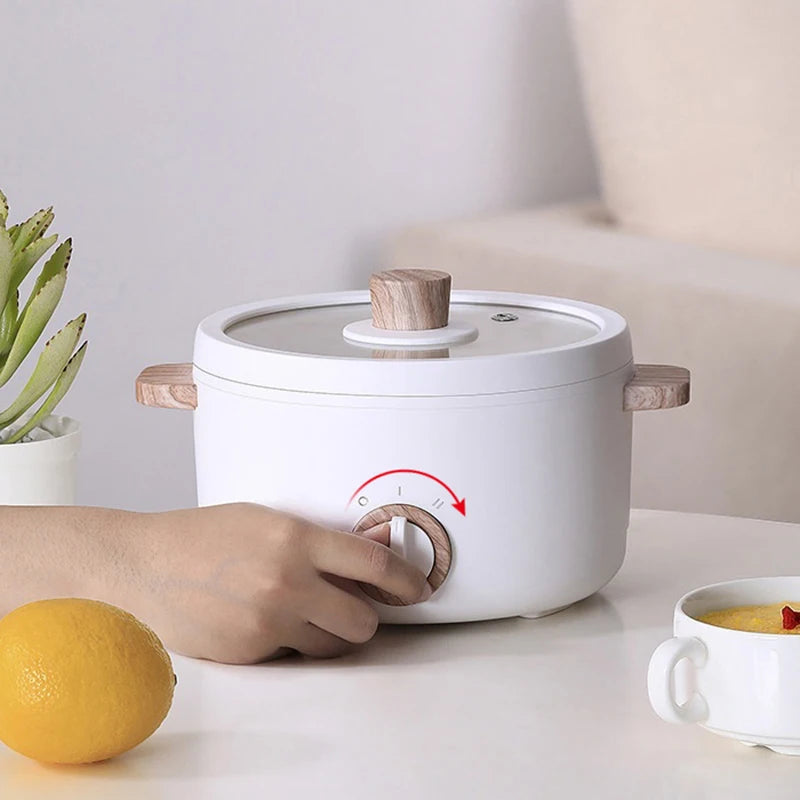Hand adjusting temperature on Multi-function Mini Cooker, set on a table with a lemon, bowl, and plant for a cozy kitchen setup.