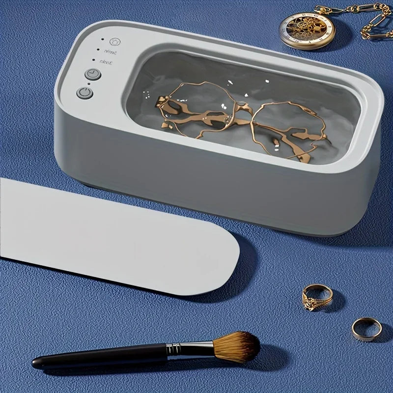 Ultrasonic cleaner in use with jewelry and makeup brush on a textured blue surface.