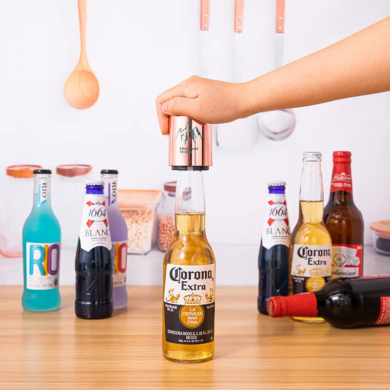 Hand using a copper can opener with mountain design to open a beer bottle on a wooden surface with other bottles around.